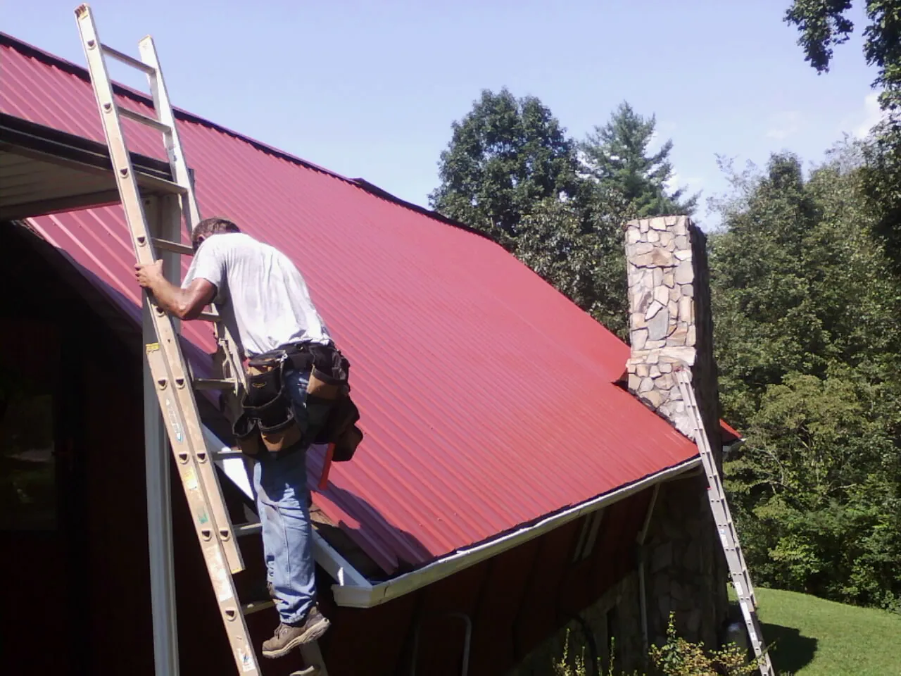 Roofer on a ladder beside a red metal roof and stone chimney — Soffit & Fascia Repair by Roofing Brothers in Gilroy